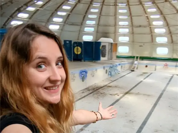Une jeune femme souriante explore une piscine tournesol abandonnée, son dôme blanc orné de fenêtres rondes.