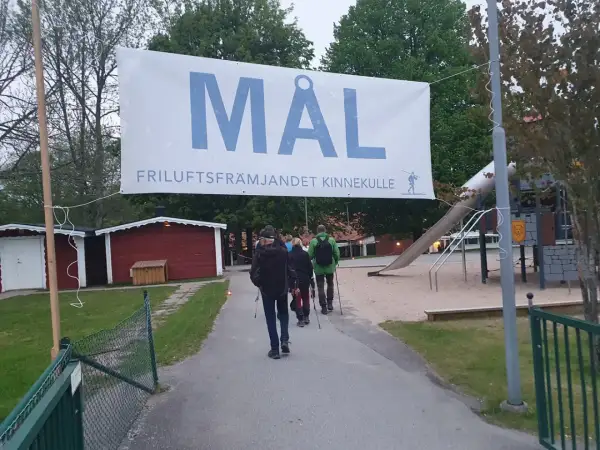 A banner reading "MÅL FRILUFTSFRÄMJANDET KINNEKULLE" hangs over a path where people walk towards a playground.