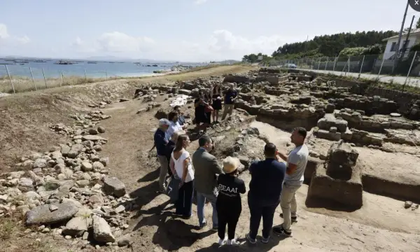 Grupo de personas observando ruinas arqueológicas en la costa. Se aprecia el mar y el **adro vello** del yacimiento.