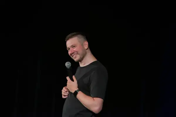 Grzegorz Wójtowicz w trakcie występu stand-up, trzyma mikrofon i uśmiecha się do publiczności.