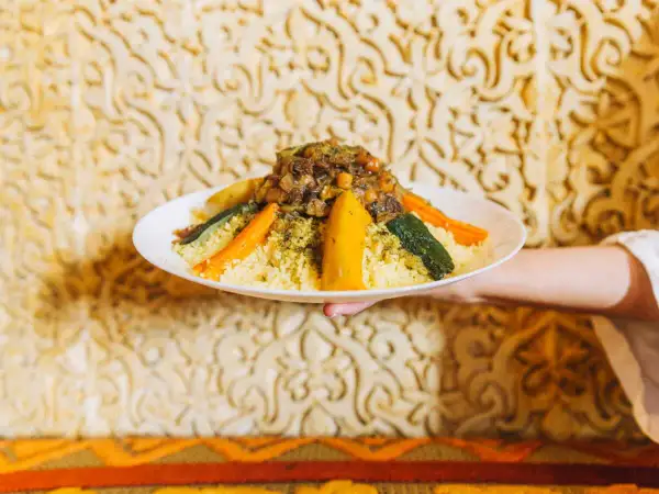 Un plat de couscous algérien, garni de légumes colorés et d'une viande savoureuse, est présenté sur un fond orné.