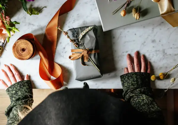 Händer som förbereder sig för att slå in runda paket med grått papper, orange sidenband och torkade blommor.