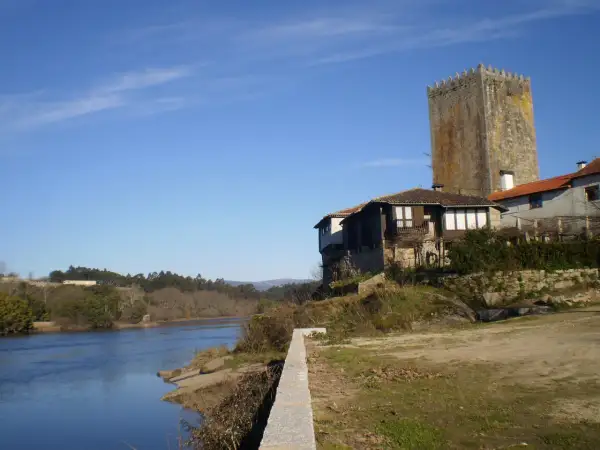 La torre de Lapela se alza junto a un río, con casas antiguas a su lado y un cielo azul despejado.