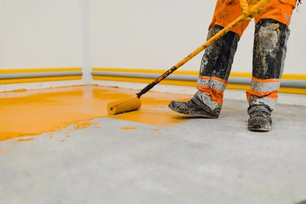 Un ouvrier en combinaison orange et noire utilise un rouleau pour peindre le sol d'un garage en jaune vif.