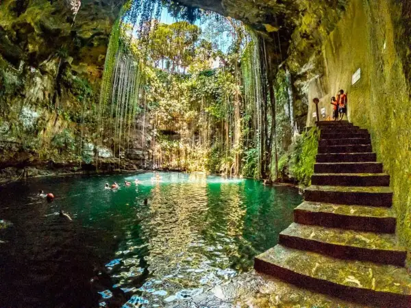 Vista interior del cenote Chichén Itzá, con escaleras de piedra y nadadores en sus aguas turquesas, rodeado de vegetación colgante.
