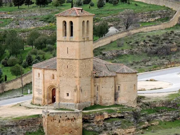La iglesia de la Veracruz, de piedra y con un campanario robusto, se alza sobre un paisaje verde y ondulado, con un camino serpenteante a su lado.