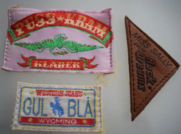 Tre märken: ett rosa med texten "PUSS & KRAM JEANS" och en grön drake, ett brunt triangelformat med "MISS SALLY FROM Rock Dreams", och ett vitt med "WESTERN JEANS GUL BLÅ WYOMING".