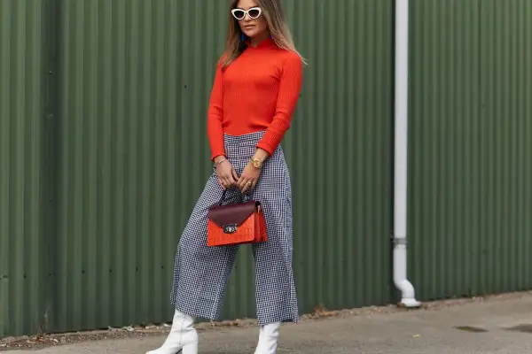Kobieta w pomarańczowym golfie i kraciastych kulotach prezentuje stylowe kuloty stylizacje. Białe kozaki i torebka dodają charakteru.