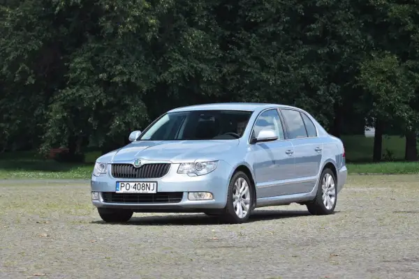 Jasnoniebieska Skoda Superb 2 z nowymi żarówkami, zaparkowana na placu przed drzewami.
