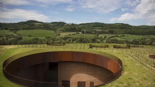 Un cerchio di metallo arrugginito emerge dal verde delle colline del Chianti, con vigneti che si estendono all'orizzonte sotto un cielo azzurro.