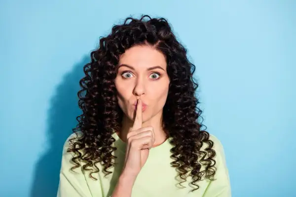 Vrouw met krullend haar houdt vinger voor de mond. Ze zegt niets, want verzwijgen is liegen.