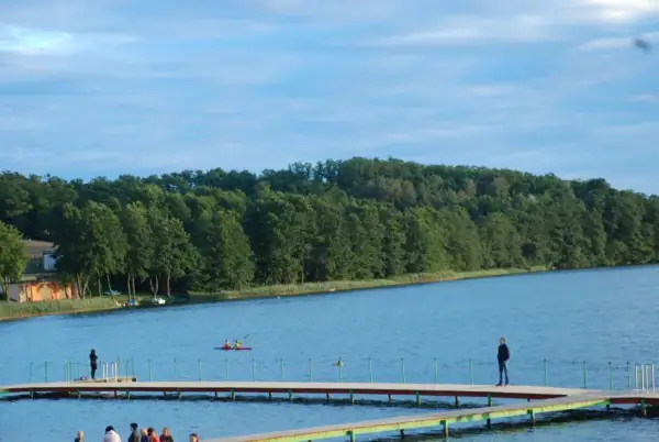 Spokojny dzień nad jeziorem Sierakowo. Ludzie spacerują po pomoście, a na wodzie pływają kajaki.