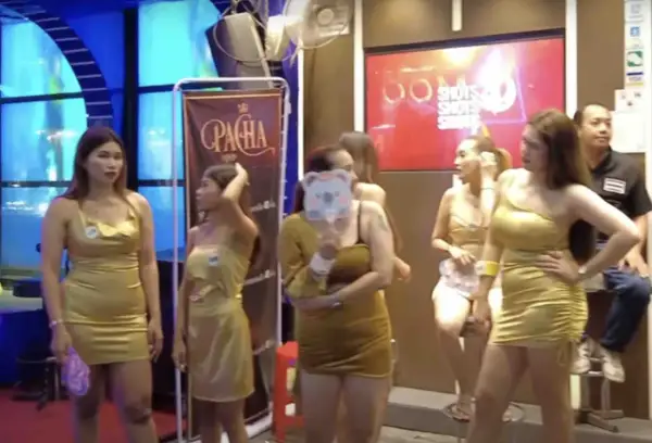 Kobiety w złotych sukienkach pozują przed banerem "Pacha". W tle neonowy napis "Shots". To scena z nocnego życia, być może związana z ping pong show w Tajlandii.