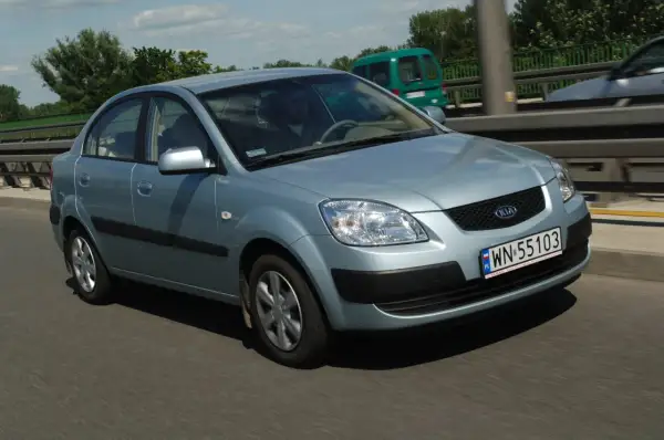 Błękitne światła Kia Rio na drodze. Kierowca w okularach prowadzi auto. W tle zielony van i drzewa.