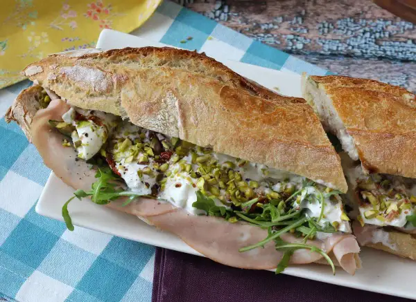 Un sandwich baguette garni de jambon, roquette, tomates séchées, pistaches concassées et burrata crémeuse.