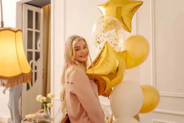 Uśmiechnięta blondynka z bukietem złotych i białych balonów w kształcie gwiazd i kuli.