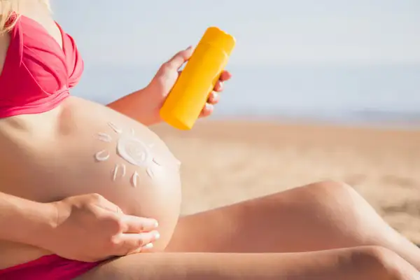 Mujer embarazada en la playa aplica protector solar en su barriga, dibujando un sol. Cuidando su piel durante el embarazo.