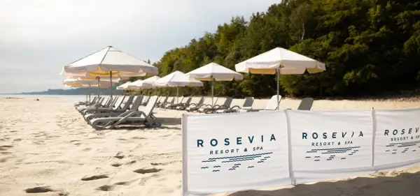 Plaża z leżakami i parasolami, w tle las, na pierwszym planie biały baner z logo "ROSEVIA RESORT & SPA".
