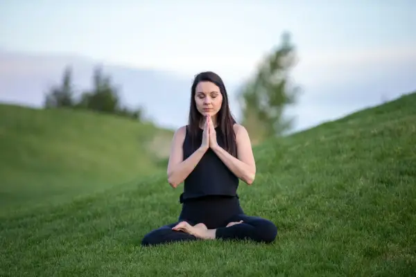 Mujer en posición de loto en la hierba, con las manos juntas en oración. Su rutina de yoga al aire libre es un momento de paz.