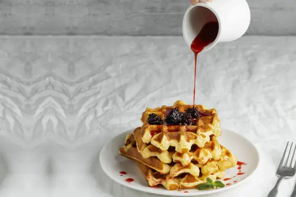 Pila di waffle dorati con more e sciroppo rosso versato da una caraffa. Ecco come fare i waffle perfetti per la colazione.
