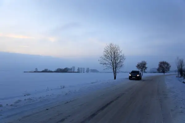Samochód jedzie po ośnieżonej drodze wśród pól. Wczesny wieczór, niebo lekko zaróżowione. Jazda po śniegu wymaga ostrożności.