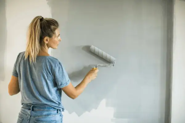 Donna con i capelli biondi in coda di cavallo, jeans e maglietta blu, impara come pitturare con il rullo una parete grigia.