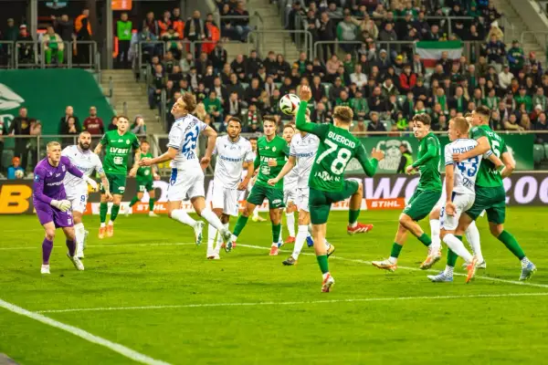 Piłkarze Lecha Poznań w białych strojach i Śląska Wrocław w zielonych grają w piłkę nożną na boisku.