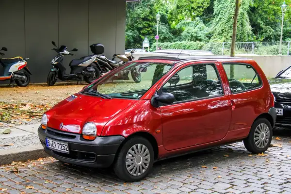 Twingo 1 rouge garée sur pavés, avec des scooters et des arbres en arrière-plan. Pensez à vérifier le filtre habitacle Twingo 1 pour un air pur.