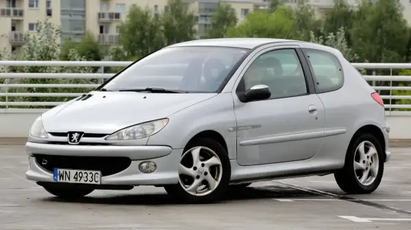 Srebrny Peugeot 206 z widocznymi przednimi żarówkami, zaparkowany na mokrym parkingu.