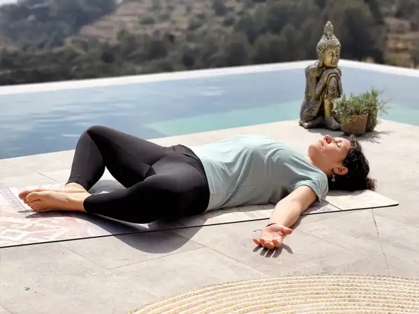 Mujer en posición de savasana, disfrutando de los beneficios de la relajación junto a una piscina, con una estatua de Buda de fondo.