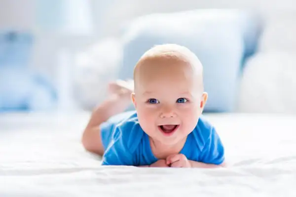 Uśmiechnięty 2 miesięczne dziecko w niebieskim body leży na brzuchu na białej pościeli.