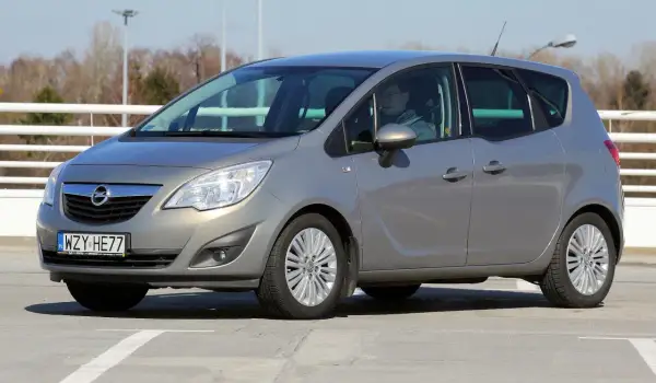 Szary Opel Meriva z kierowcą za kierownicą, zaparkowany na betonowym placu.