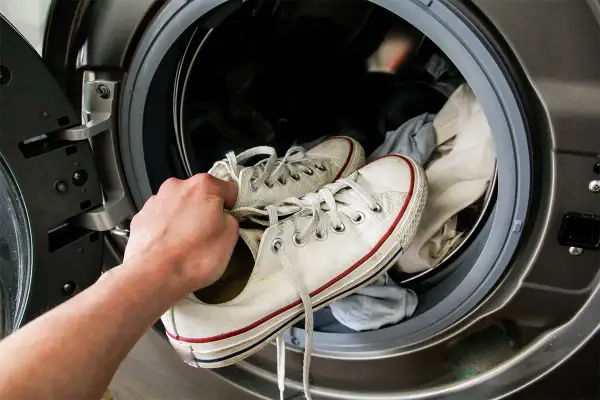 Mano che inserisce scarpe da ginnastica bianche in lavatrice per lavarle. La centrifuga le renderà come nuove!