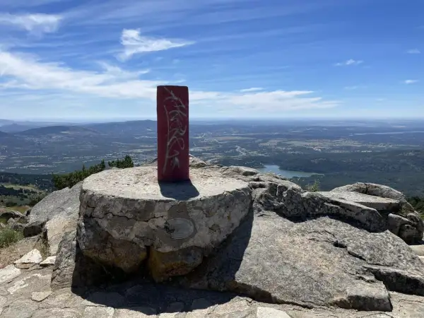 Pilar rojo con grafitis en la cima de una montaña, con un paisaje de valle y lago al fondo. La **cabeza lijar** del pilar parece desgastada por el tiempo.