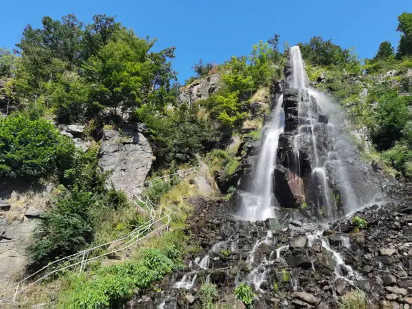 Der Trusetaler Wasserfall stürzt über schwarze Felsen in die Tiefe. Ein weißer Geländerweg führt Besucher am grünen Hang entlang.