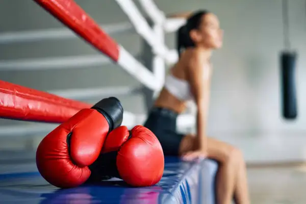 Red boxing gloves rest on the edge of a boxing ring. A woman sits in the background, perhaps contemplating how to do a body shot.