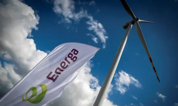 Flaga z logo Energa powiewa na tle błękitnego nieba z chmurami, obok wiatraka. Akcje Energa rosną.