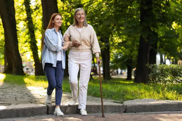 Młoda kobieta pomaga starszej pani, która wspiera się na czwórnodze, spacerując po parku.
