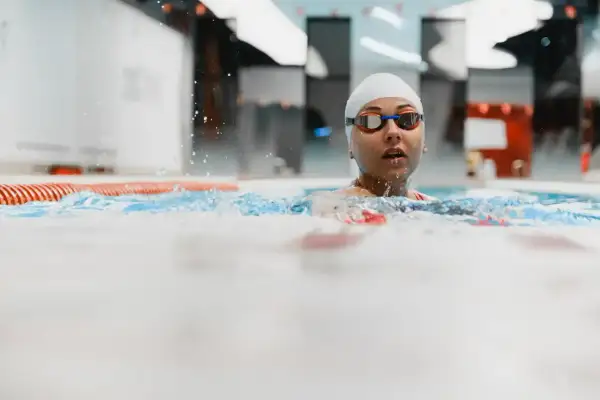 Kobieta w czepku i okularach pływackich w basenie, z wodą rozpryskującą się wokół.
