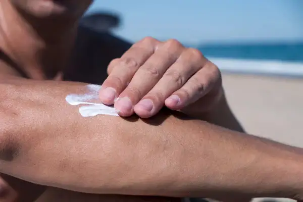 Dłoń rozsmarowuje krem z filtrem, zawierający m.in. diethylamino hydroxybenzoyl hexyl benzoate, na ramieniu na plaży.