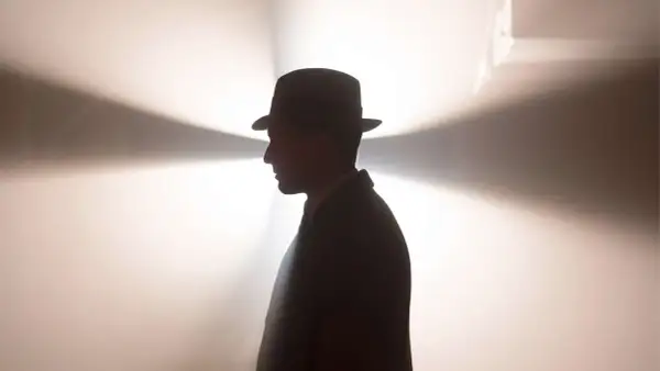 Ein Mann im Hut und Anzug, im Profil, vor einem hellen, strahlenden Hintergrund. Ein mysteriöser Blick, der an Babylon Berlin Staffel 5 erinnert.