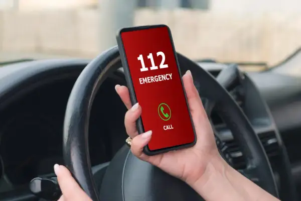 Mano sujetando un teléfono con pantalla roja que muestra "112 EMERGENCY" y un botón de llamada verde. El volante del coche está visible.
