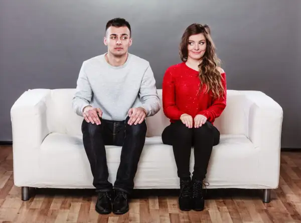 Coppia seduta su un divano bianco, lui guarda altrove con espressione incerta, lei sorride. Come superare la timidezza in amore?