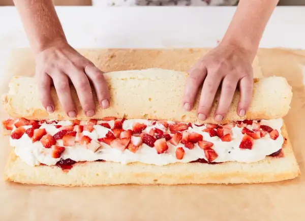 Hände rollen einen Biskuitteig mit Sahne und Erdbeeren für ein Erdbeerrolle Rezept auf.