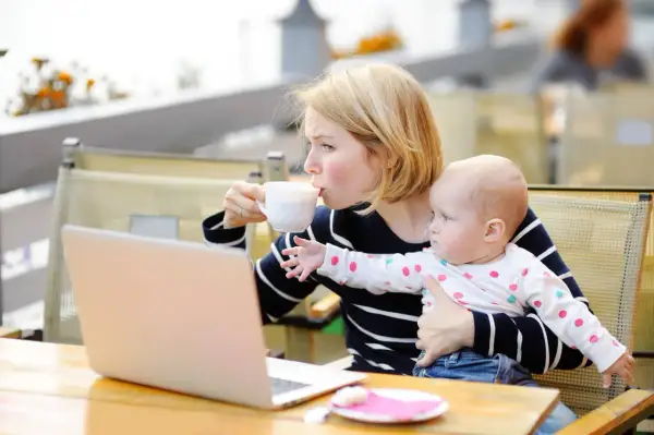 Kobieta pije kawę, trzymając dziecko. Obok leży laptop. Karmienie piersią i chwila dla siebie.