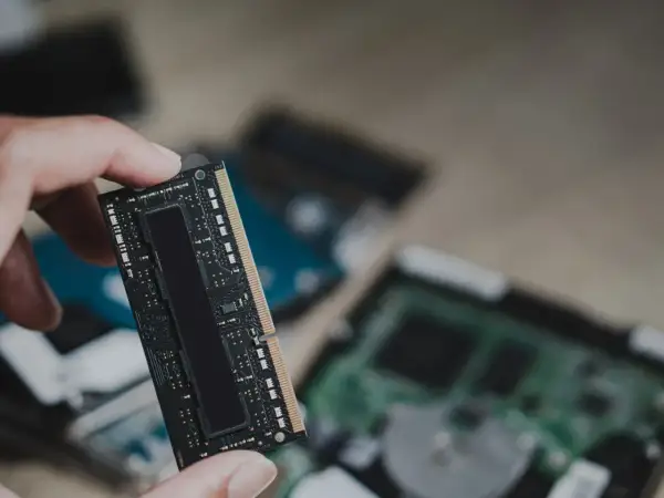 Dłoń trzyma moduł pamięci RAM, prawdopodobnie LPDDR5, z widocznymi układami scalonymi. W tle widać inne komponenty komputerowe.