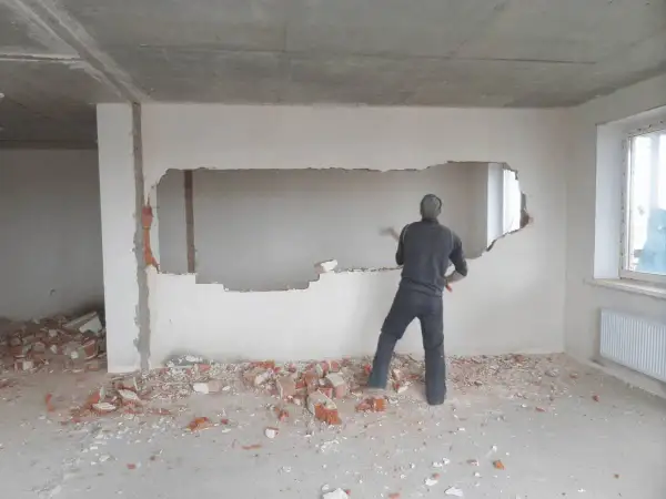 Mężczyzna z młotem wyburza fragment ściany nośnej, wokół gruz.