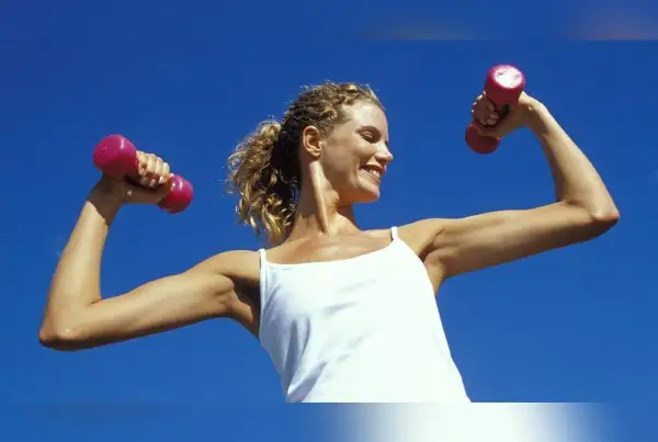 Femme souriante avec des haltères roses, montrant ses muscle bras. Elle porte un débardeur blanc sous un ciel bleu clair.