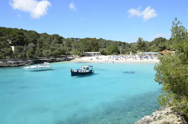 En solig dag på mallis Mallorca. Turkosblått vatten, en vit yacht och en blåvit båt i en vik med en livlig strand.