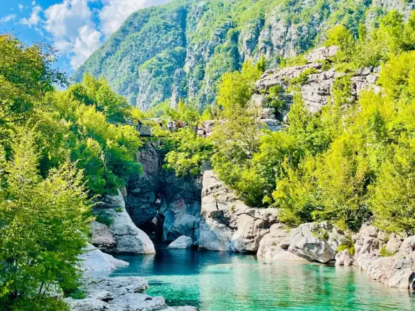Aguas turquesas fluyen entre rocas y vegetación exuberante, bajo imponentes montañas en Albania. Un puente peatonal cruza el cañón, invitando a explorar las **montañas malditas de Albania**.